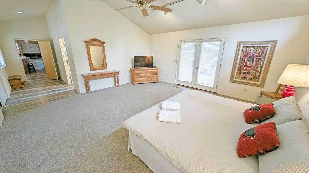 A king-sized bed takes center stage in a spacious bedroom, complemented by elegant wall art, French doors flooding the space with sunlight and providing access to the outside, and a television atop a dresser. The room feels relaxing and connected to nature.