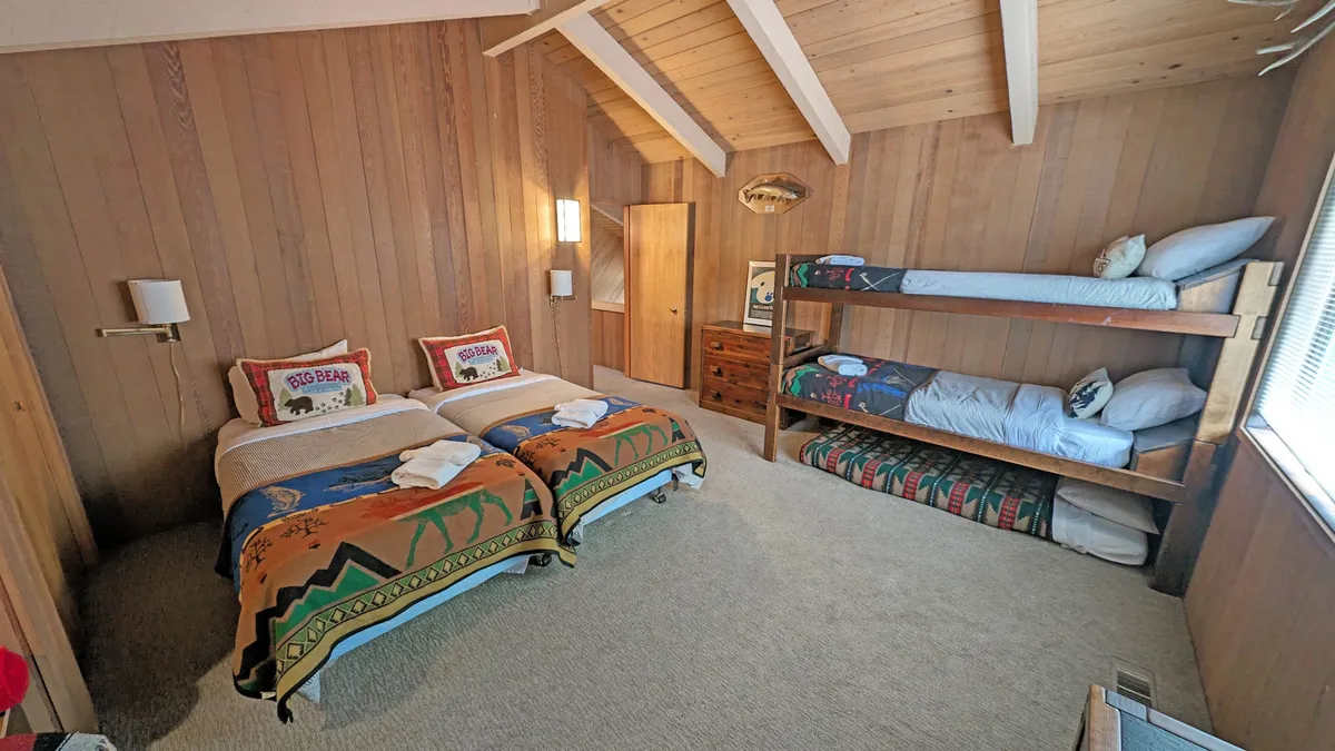 A playful upstairs kids’ bedroom featuring two twin beds and a bunk bed with colorful, decorative blankets and wooden paneling. The room exudes warmth and provides an imaginative retreat for younger guests, perfect for reading, playing, and relaxing.