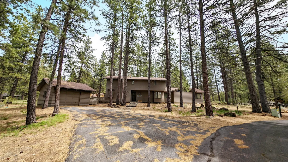 This exterior image showcases the house nestled among towering pine trees, with a driveway leading up to the entrance. The peaceful surroundings and ample outdoor space suggest the home is situated in a natural, serene setting ideal for family gatherings and exploration.
