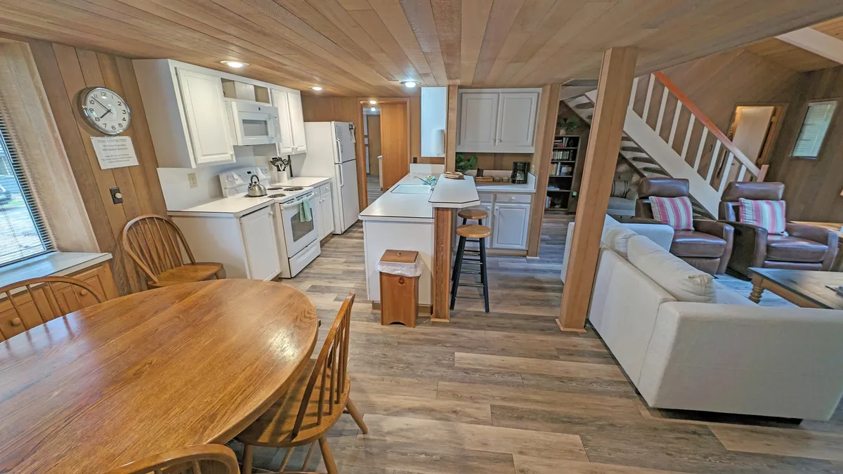 An integrated space featuring a dining table, kitchen island, and adjacent living room seating, all illuminated by warm lighting. The scene exudes a welcoming and communal atmosphere, perfect for group activities and shared meals.