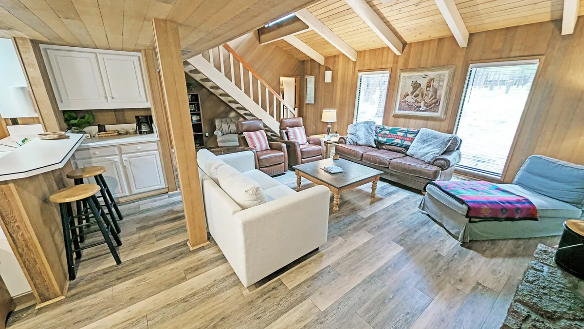 The image showcases a bright and inviting living room with plush sofas arranged around a modern coffee table, a striking staircase, and expansive windows that flood the space with natural light. The setup is perfect for relaxing, socializing, or gathering for games and conversation, all within a cozy, welcoming atmosphere.