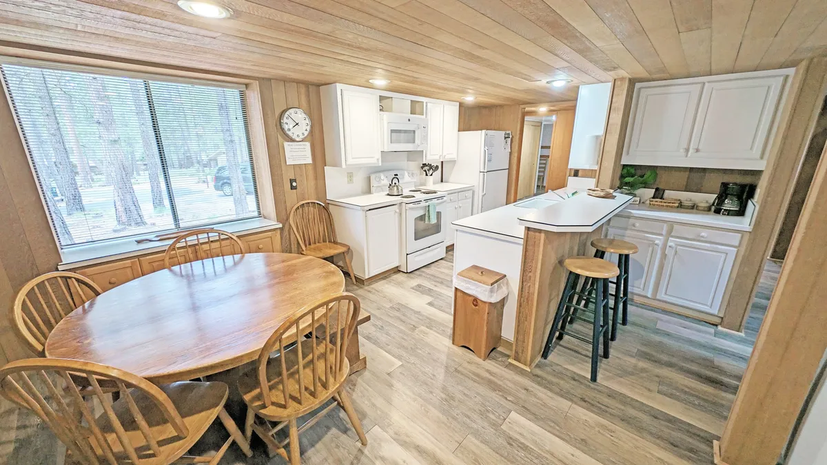 A bright dining area with a round wooden table set near a large window, white cabinets in the background, and beautiful natural light streaming in. The setup is ideal for intimate family meals and casual gatherings.