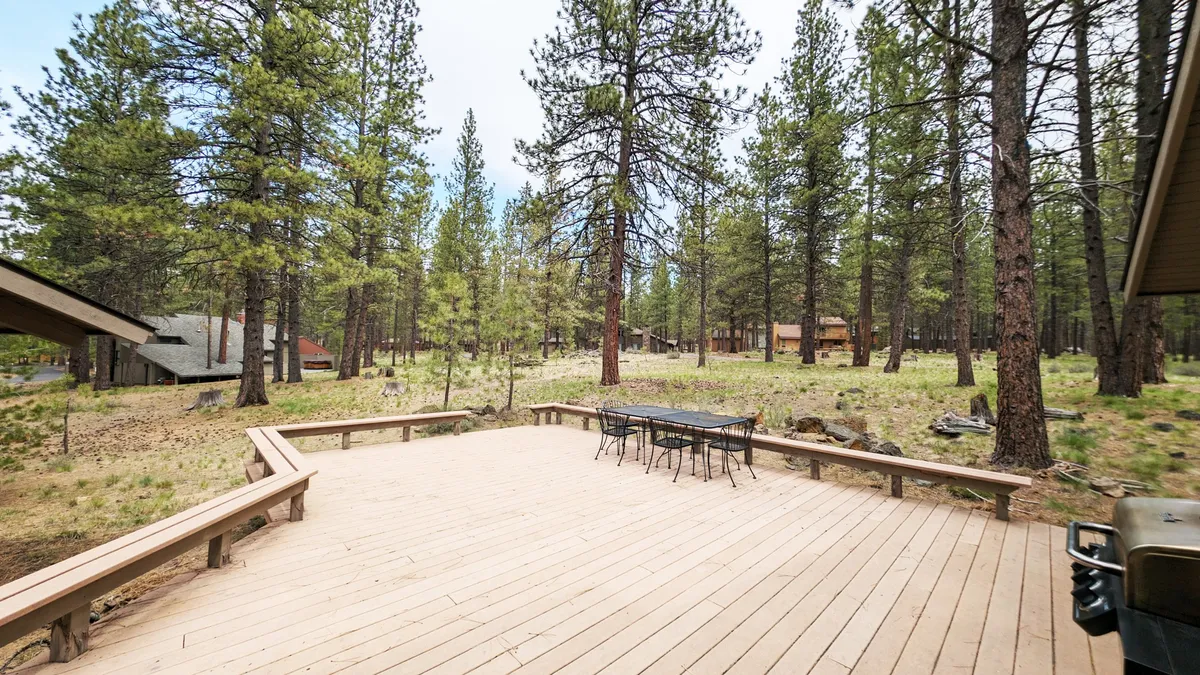 A spacious wooden deck surrounded by tall trees, featuring an outdoor dining table set up for meals and relaxation. Natural sunlight filters through the foliage, creating a tranquil, private oasis ideal for gatherings or quiet moments.