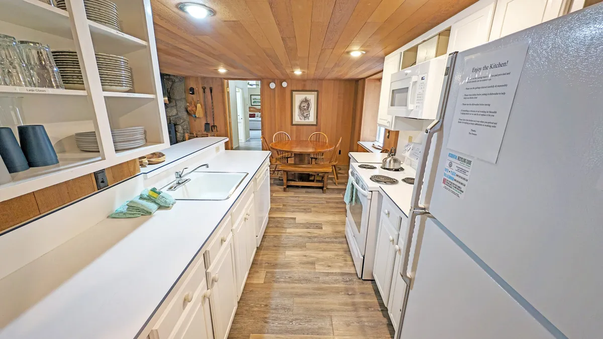 This image focuses on a cozy kitchen and dining space, with a wooden dining table prominently set near a light-filled kitchen countertop and white cabinetry. The inviting wood ceiling and open space suggest a setting ready for family feasts and group gatherings.