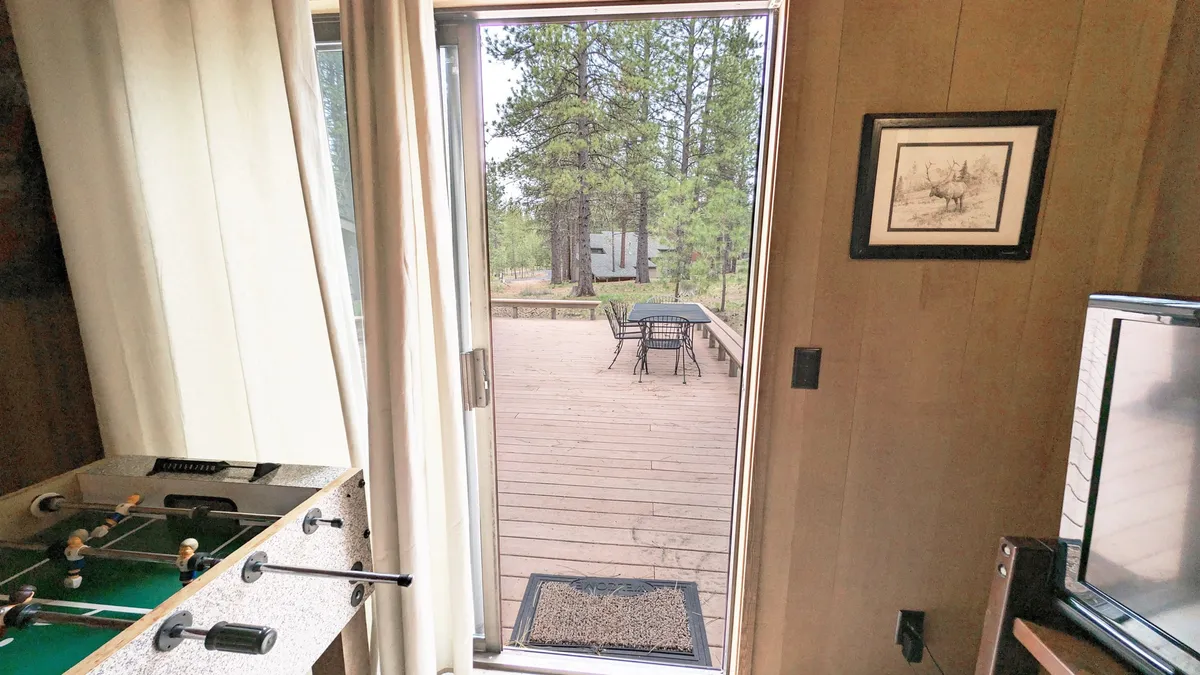 A game room equipped with a foosball table, large doorway opening onto the deck, and an outdoor dining area visible just beyond the glass. The space is designed for both fun and easy transitions between indoor and outdoor activities.