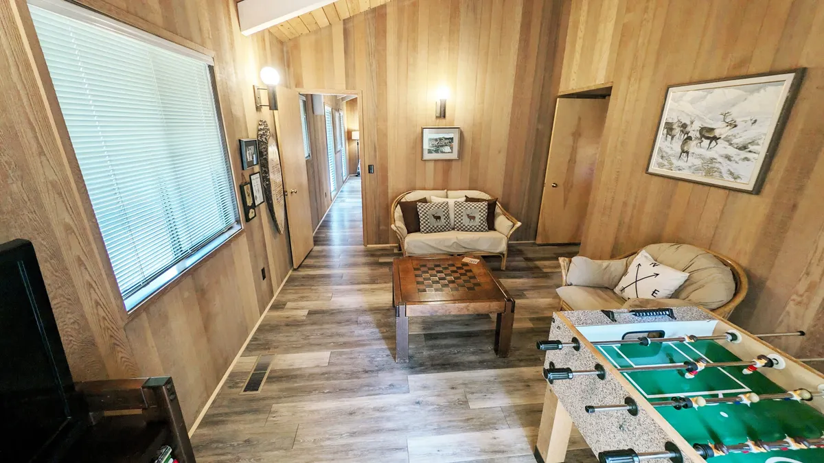 This image captures the heart of the Den/game room, featuring a foosball table and a checkerboard table at the center, surrounded by a sofa with colorful cushions and bright, playful artwork on the wall. The mood is casual and friendly, with space for both competitive games and relaxed socializing.